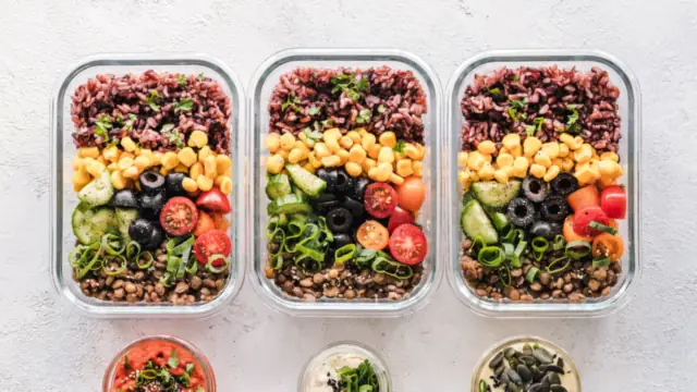 Three glass meal prep containers packed with rice, corn, olives, tomatoes and lentils, with small jars of sauce and seeds