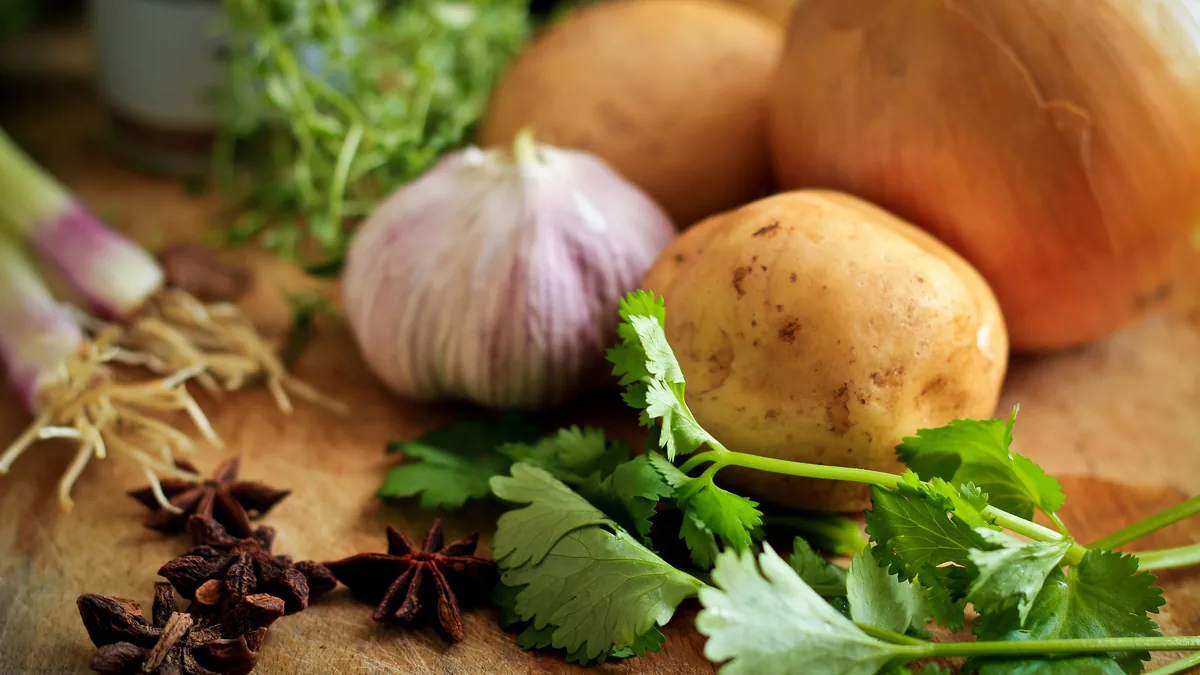 Low-cost staples on a wooden board: potatoes, an onion, garlic, fresh cilantro and whole spices